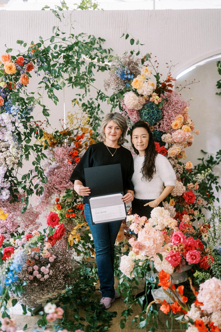 Two women standing among vibrant floral arrangements, holding a certificate folder. The background is filled with a variety of colorful flowers and lush greenery, creating an elegant and celebratory atmosphere.