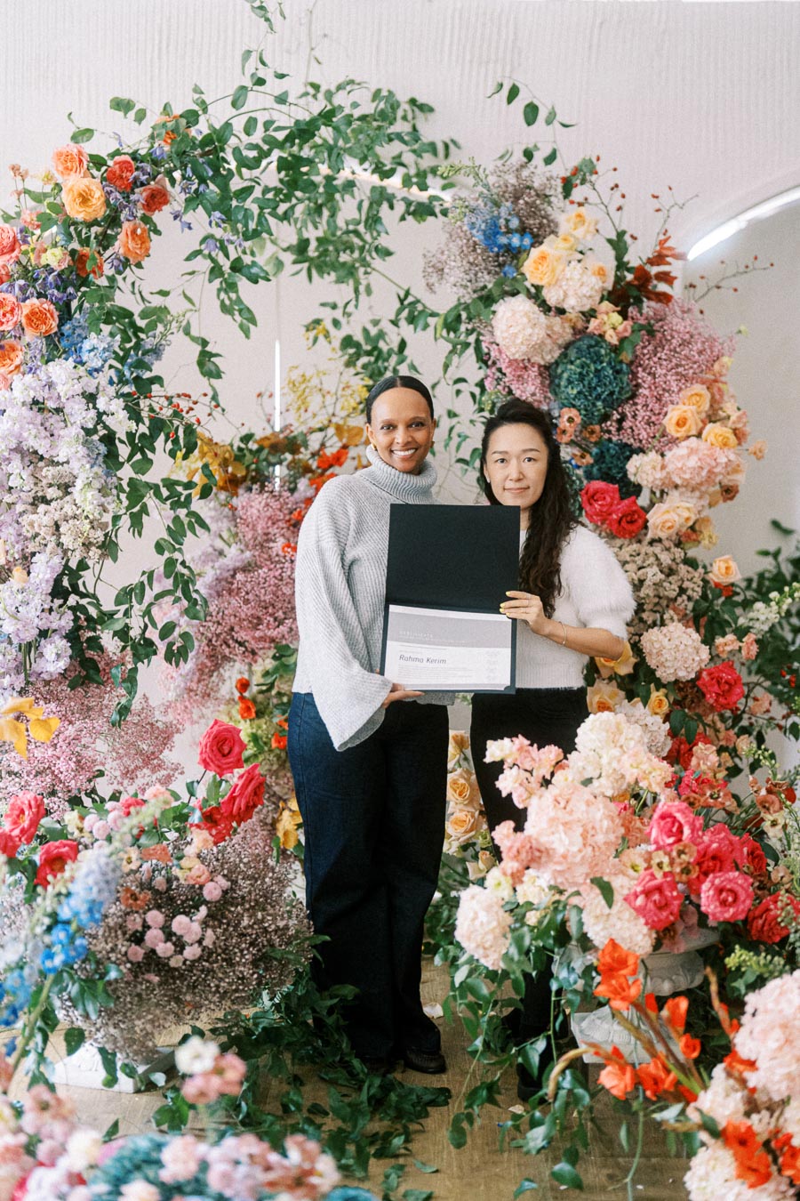 Two people smiling and holding a certificate, surrounded by a vibrant floral arrangement featuring roses, hydrangeas, and greenery, creating an elegant and celebratory atmosphere.