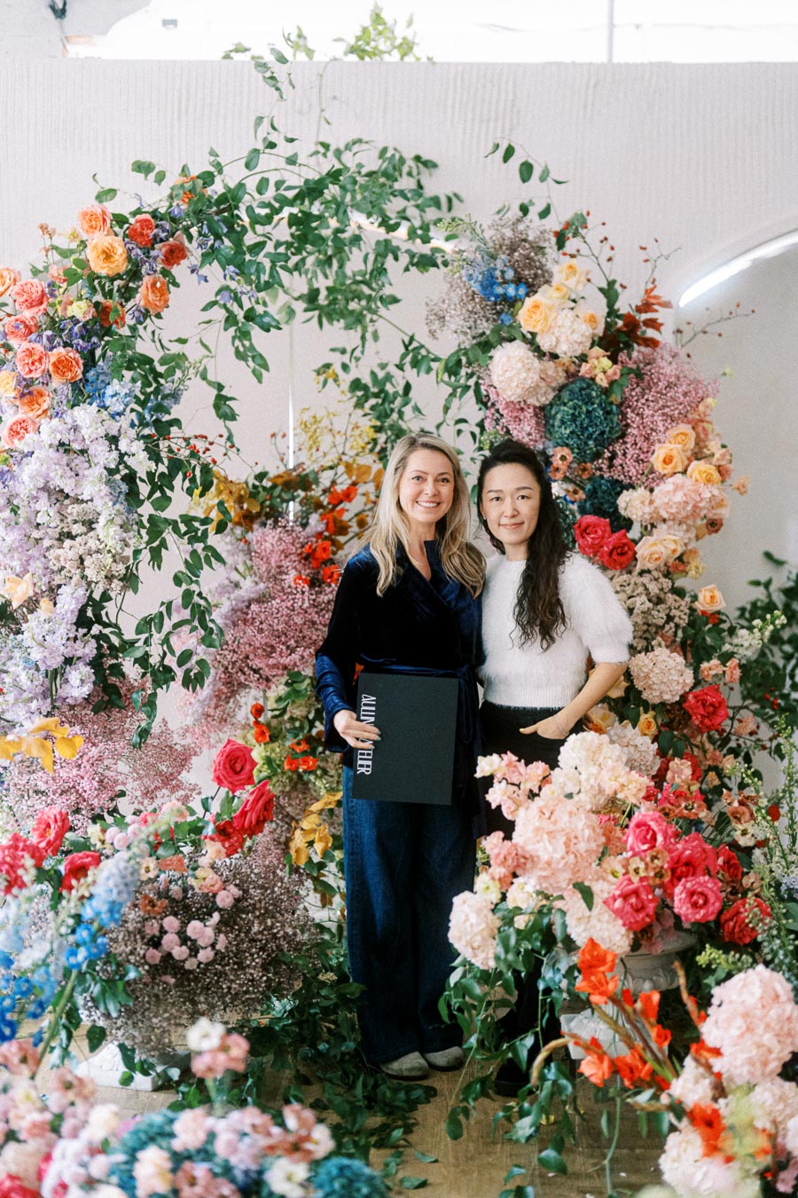 Two people smiling and posing in front of a vibrant floral display featuring a variety of colorful flowers and lush greenery. One person is holding a black book titled Album Fever.