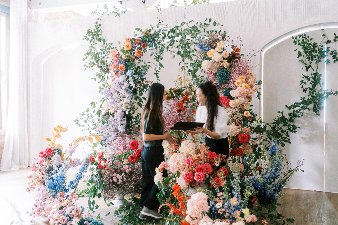 Two women interacting in a beautifully decorated flower shop, surrounded by vibrant floral arrangements featuring roses, lilacs, and greenery against a white background.