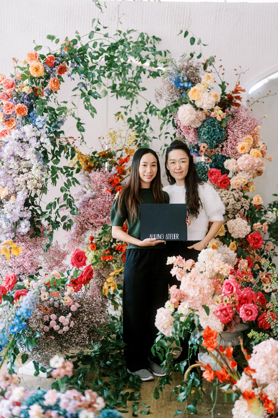 Two people standing in front of a vibrant floral arrangement, holding a sign that reads Allung Atelier. The display features an array of colorful flowers, including roses and hydrangeas, creating a lush, decorative backdrop.