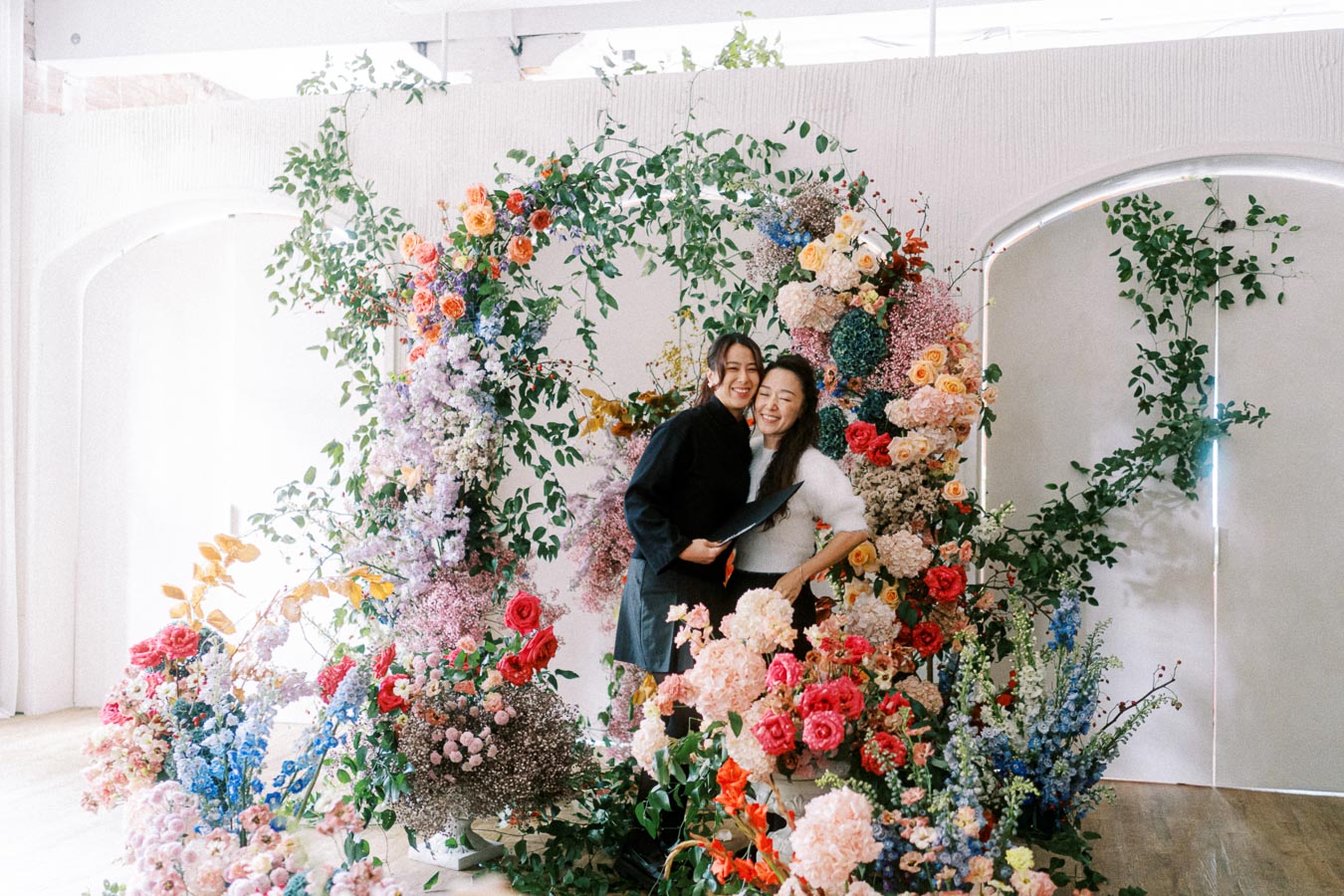 Two people smiling joyfully in front of a vibrant, colorful floral arrangement featuring roses, hydrangeas, and greenery, indoors with a light background.