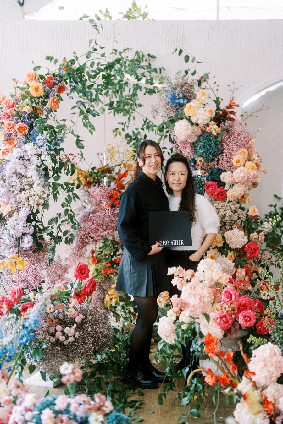 Two people standing together among a vibrant floral display, showcasing colorful arrangements of roses, hydrangeas, and greenery indoors. One person holds a black folder with text, creating an elegant and artistic setting. Ideal for a celebration or event highlight featuring floral design and teamwork.