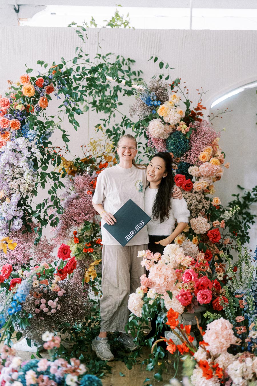 Two individuals standing happily in front of a vibrant floral arch, showcasing a diverse array of colorful flowers including roses and hydrangeas. They are holding a folder labeled Ailing Aher, capturing a cheerful and creative moment in a floral exhibit or workshop.