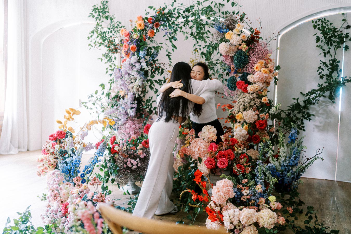 Two people embracing warmly surrounded by a lush, colorful floral arrangement with vibrant blooms and greenery in a bright, airy space.