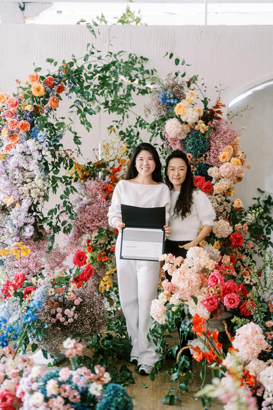 Two women smiling and standing in front of a colorful, lush floral arrangement, with one holding a certificate. Perfect for springtime events or floral design themes.