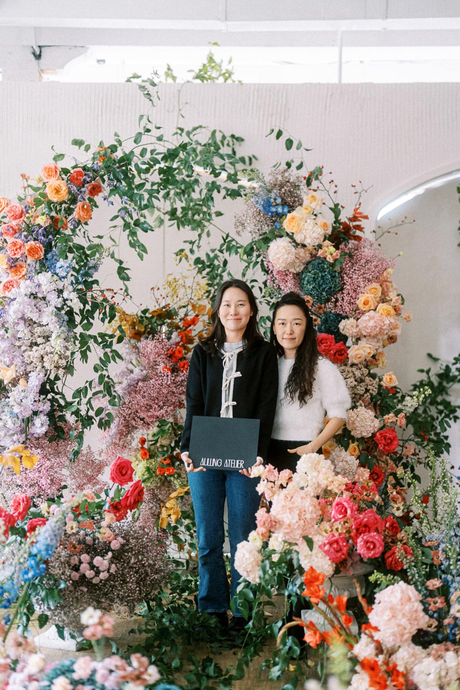 Two women standing amidst a vibrant floral arrangement, holding a sign reading Allung Atelier. Surrounded by colorful flowers and greenery, creating a lush, artistic display.