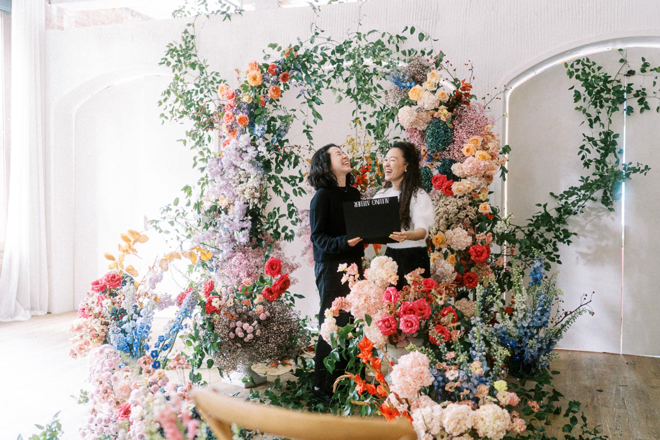 Two people joyfully stand surrounded by a vibrant floral arrangement, holding a black box, suggesting celebration and elegance.