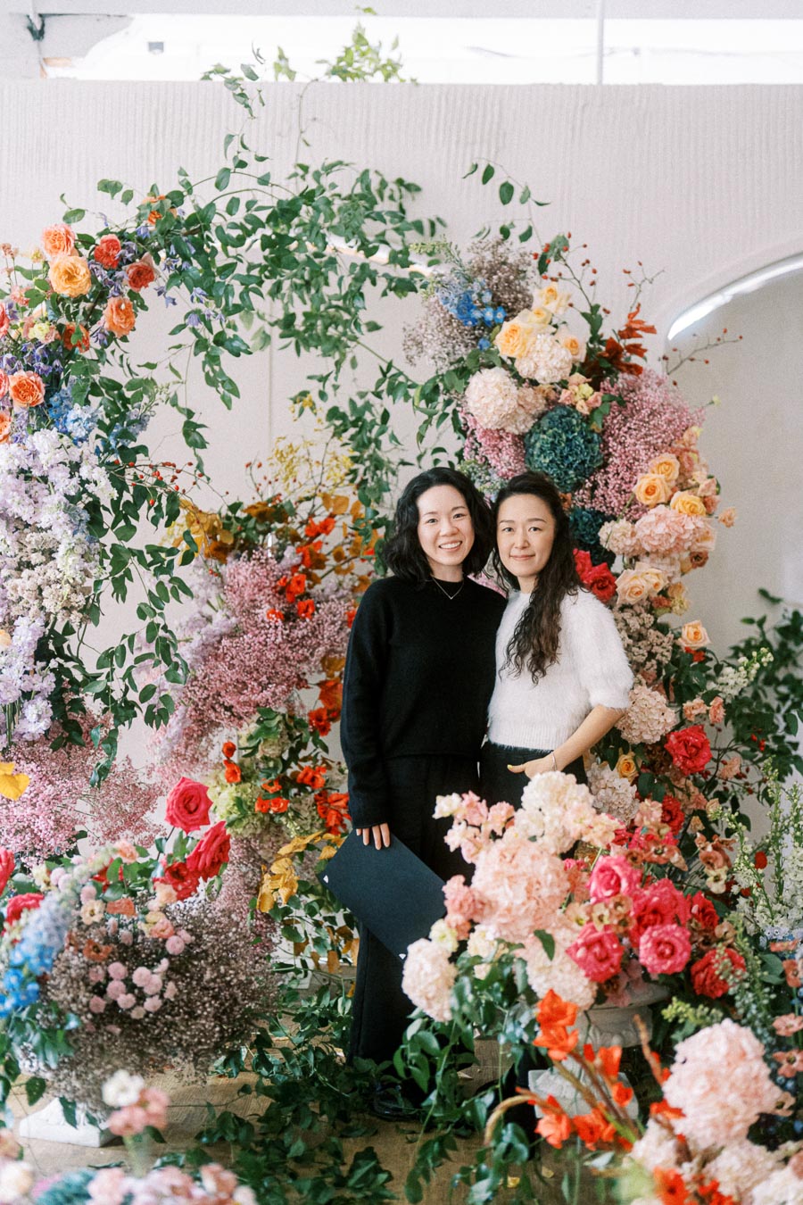 Two individuals stand smiling in front of a vibrant floral arrangement featuring an array of colorful blooms including roses and hydrangeas, alongside lush green foliage. The background showcases a soft, elegant garden-themed setting perfect for a festive or celebratory occasion.
