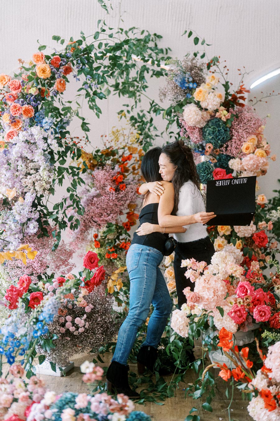 Two women embrace joyfully amidst a lush, colorful floral arrangement, featuring a variety of vibrant flowers and greenery, as one holds a black box.