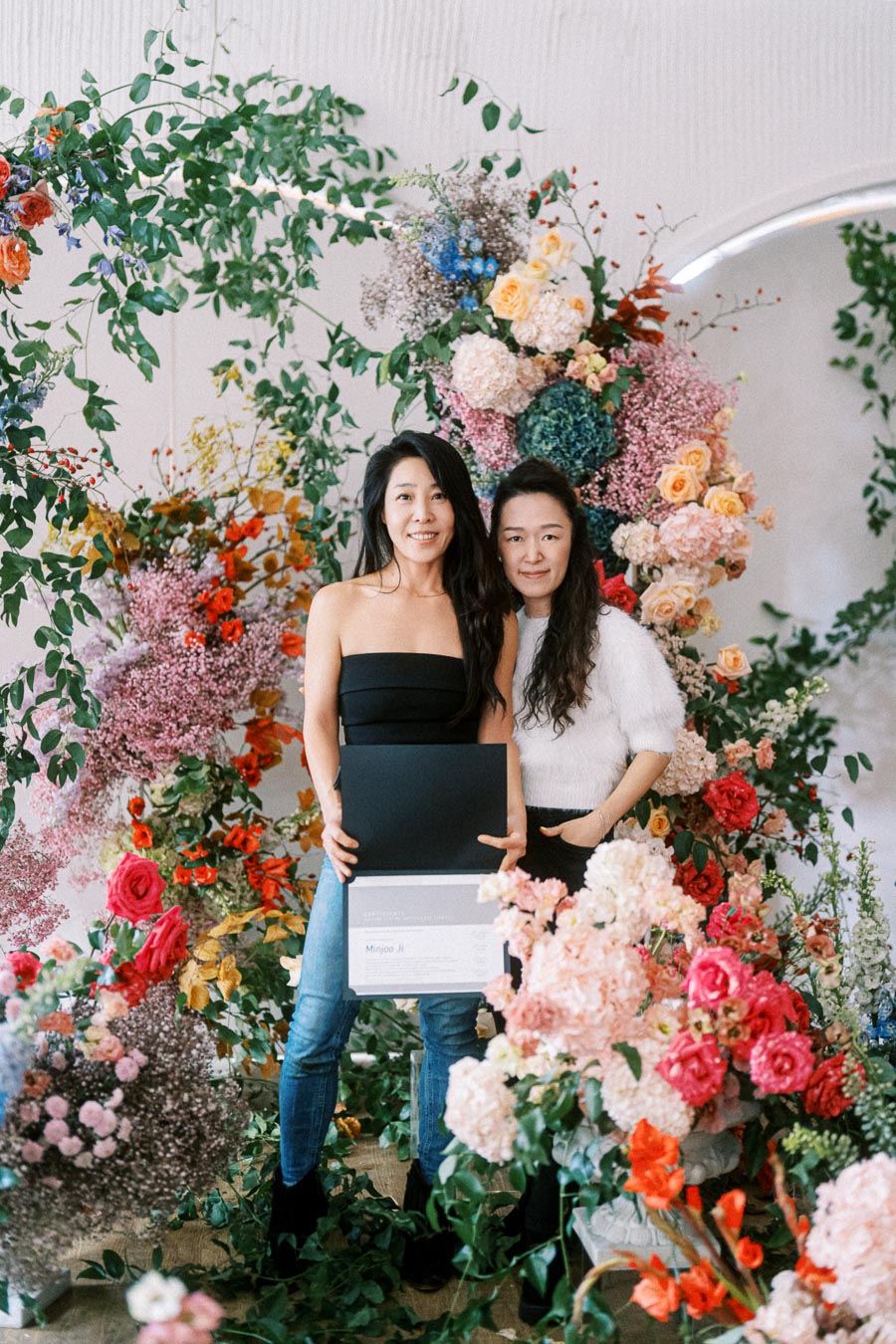 Two women standing in front of an elaborate floral arrangement, holding a certificate. The vibrant display features roses, hydrangeas, and lush greenery, creating a colorful and celebratory setting.