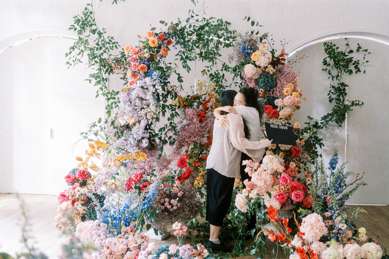 A person holding a bouquet of vibrant, colorful flowers stands next to a lush, artistic floral display featuring roses, peonies, and greenery inside a bright room with arched doorways.