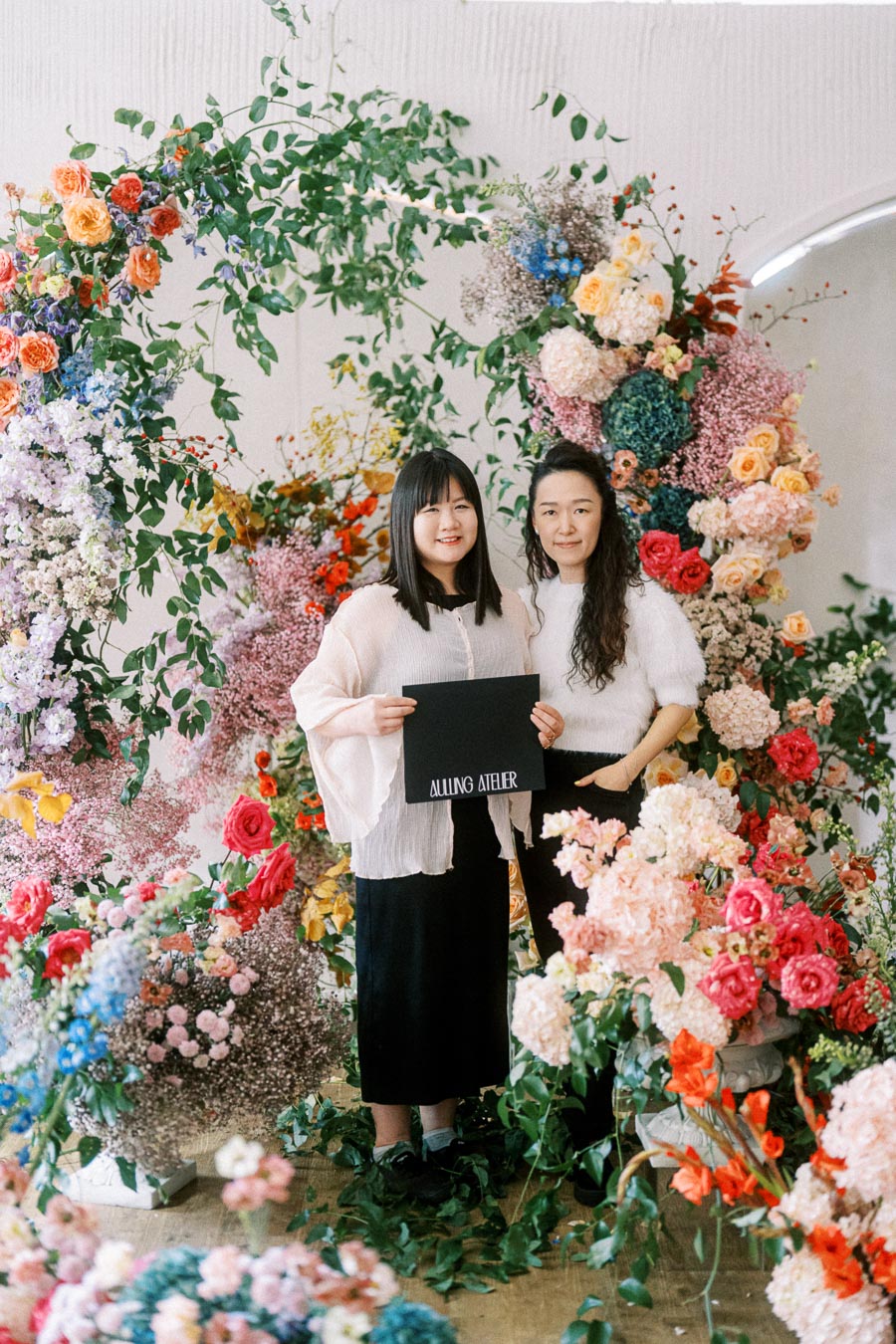 Two women standing amidst a vibrant, colorful floral arrangement, holding a sign that reads Allung Atelier, showcasing a creative floral design exhibition.