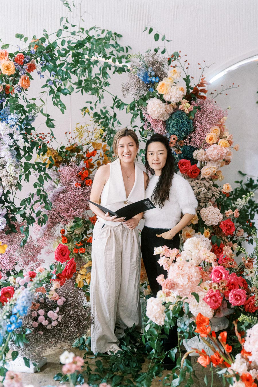 Two women standing in front of a vibrant floral arrangement featuring colorful roses, hydrangeas, and greenery, creating a lush backdrop.