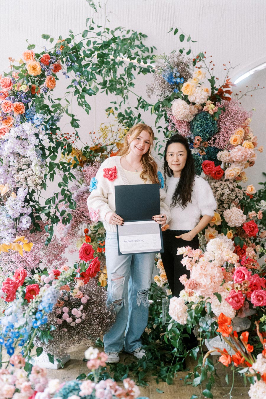 Two women smiling while standing amidst a lush floral arrangement with vibrant roses, hydrangeas, and greenery, one holding a certificate in her hands.