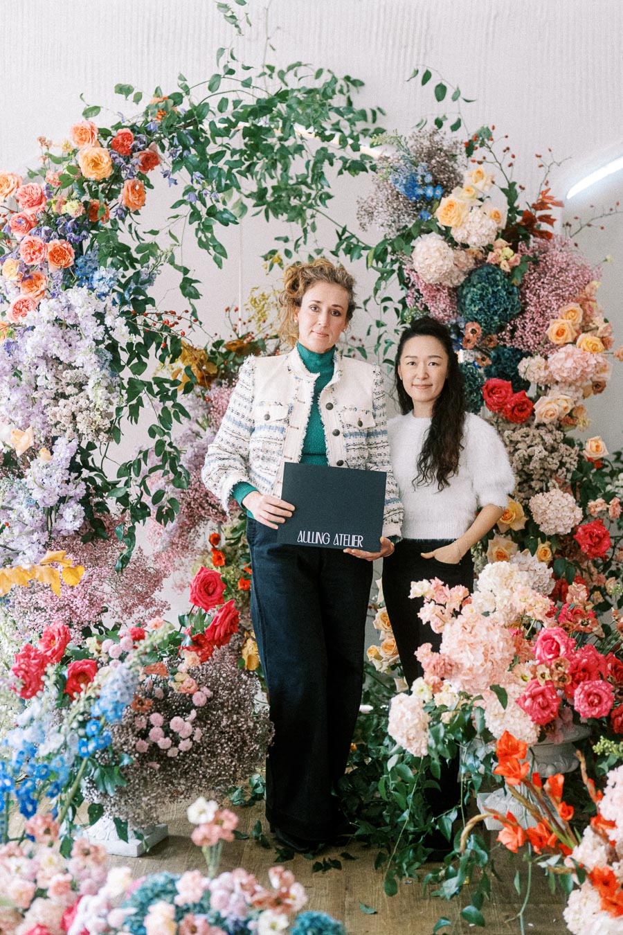 Two women stand amidst a vibrant floral arrangement holding a sign that reads Ailing Atelier. The scene is filled with colorful roses, hydrangeas, and lush greenery, creating a lively and artistic backdrop.