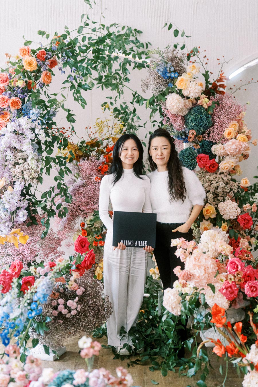 Two women stand surrounded by a vibrant display of colorful flowers and greenery, holding a sign that reads Falling Atelier. They are dressed in coordinated white outfits.