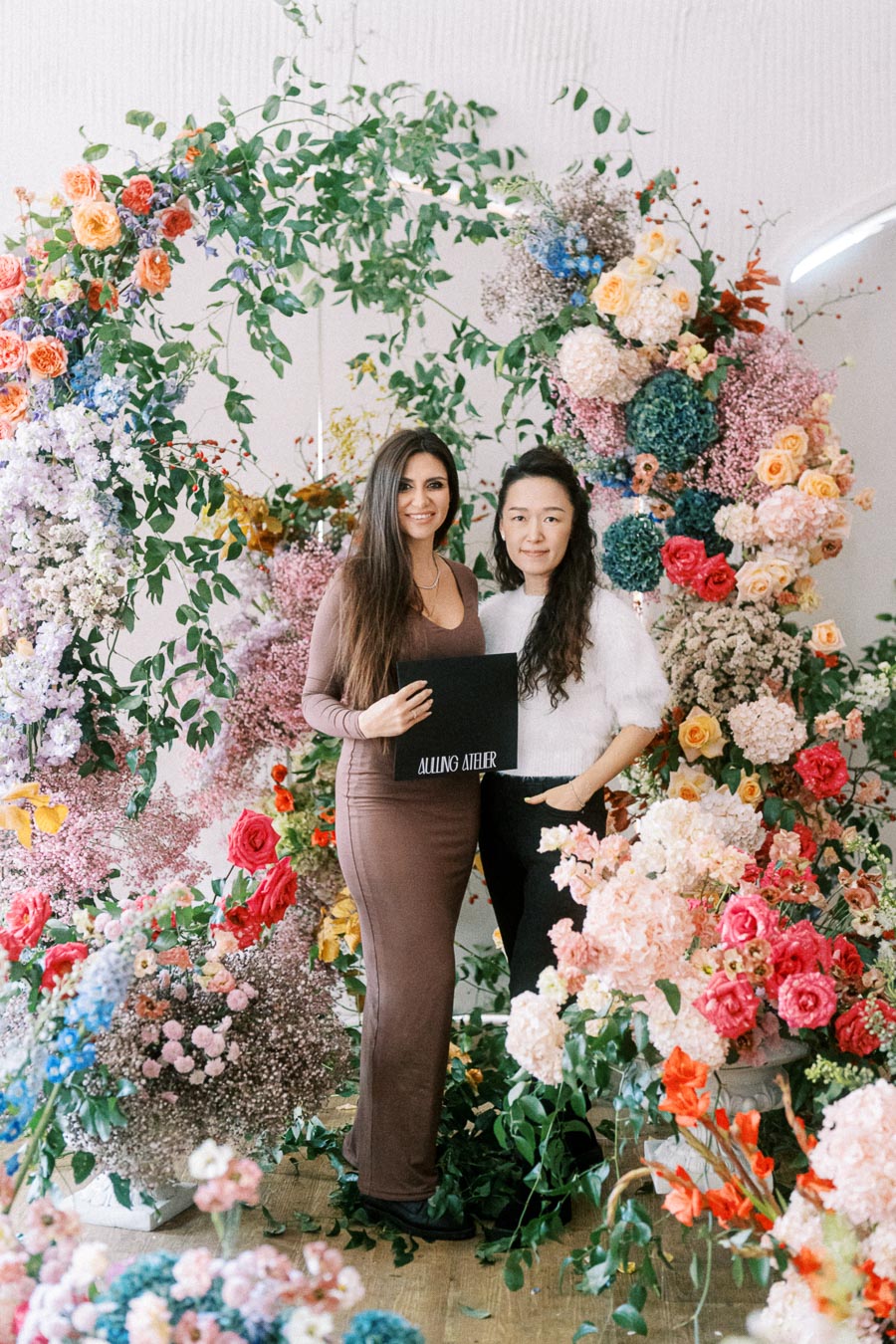 Two women standing in front of an elaborate floral arrangement featuring a variety of colorful flowers and green foliage. One woman is holding a black folder, and they are both smiling. The background showcases vibrant colors with pink, red, blue, and orange flowers.