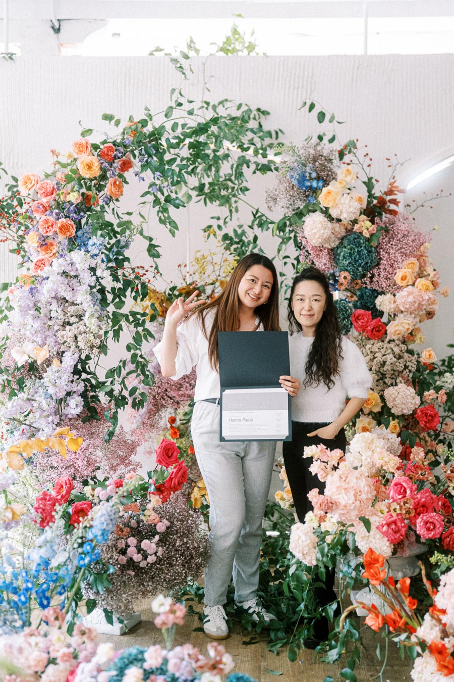 Two women smiling and posing in front of an elaborate floral arch, showcasing a certificate. The scene is vibrant with a variety of colorful flowers including roses, hydrangeas, and greenery, creating a lush and celebratory atmosphere.