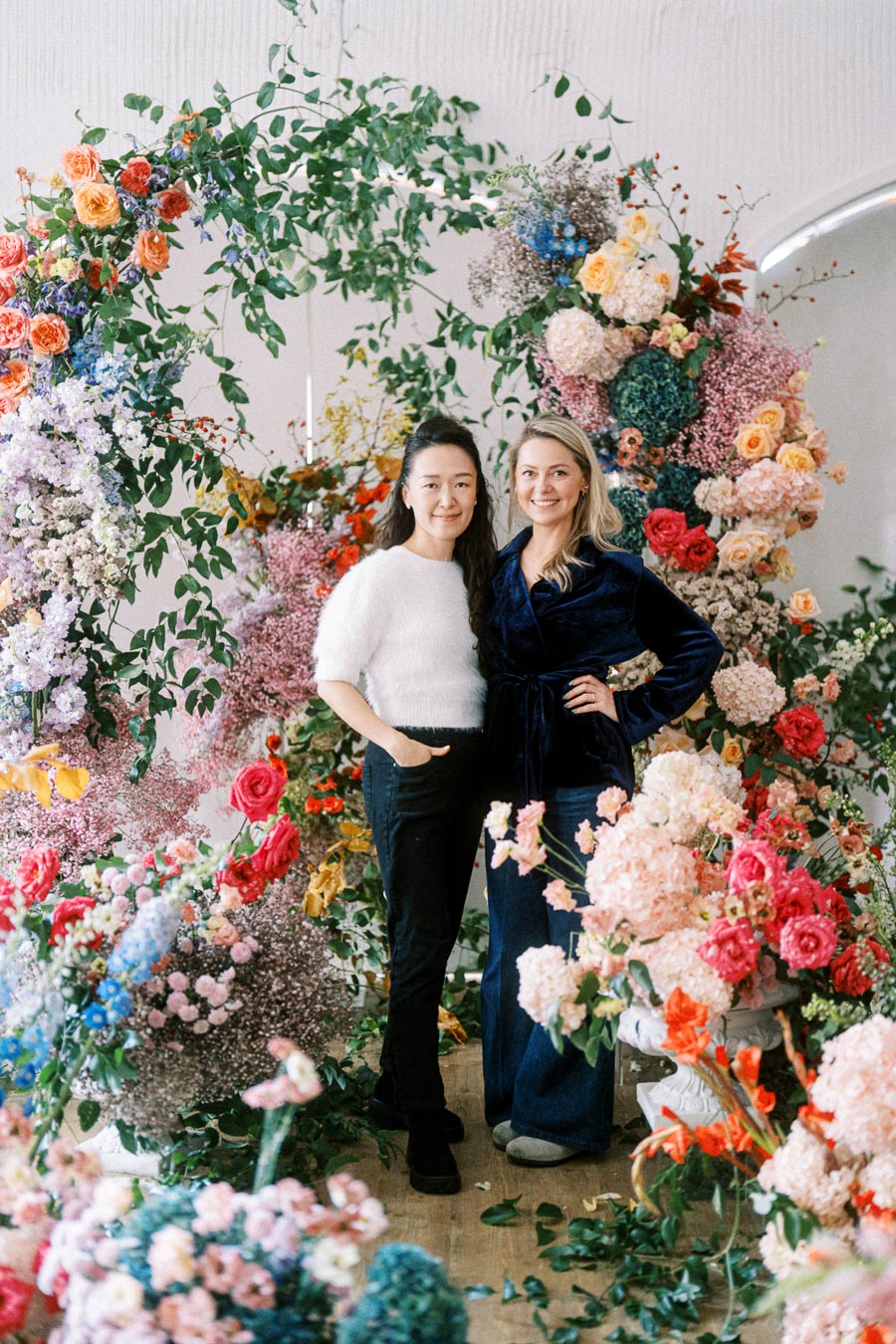 Two people standing amidst a vibrant floral arch, featuring an array of colorful blooms including roses, hydrangeas, and greenery. The setting is indoors, radiating a celebratory and artistic atmosphere.