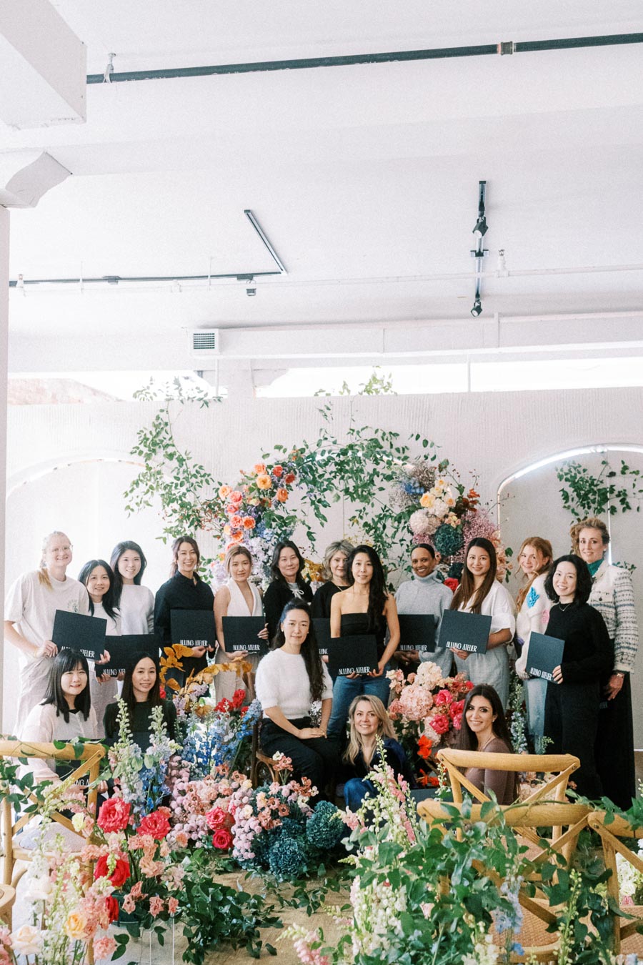 A diverse group of women holding certificates, posing amidst vibrant floral arrangements and greenery in a well-lit room, suggesting a graduation or workshop event focused on floral design.