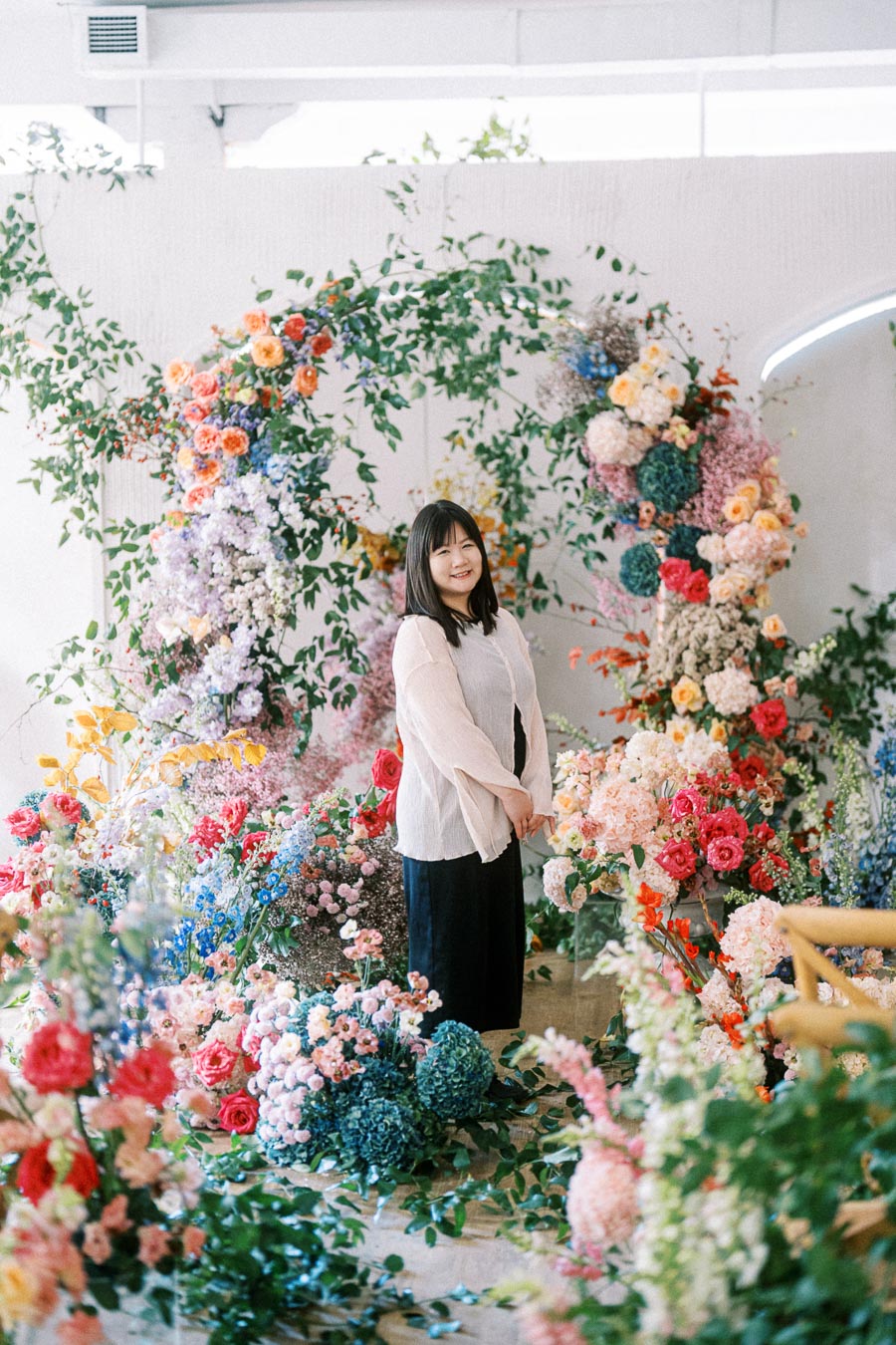 A person standing amidst a vibrant floral arrangement, featuring a circular arch decorated with various colorful flowers including roses and hydrangeas, in a bright, indoor setting.
