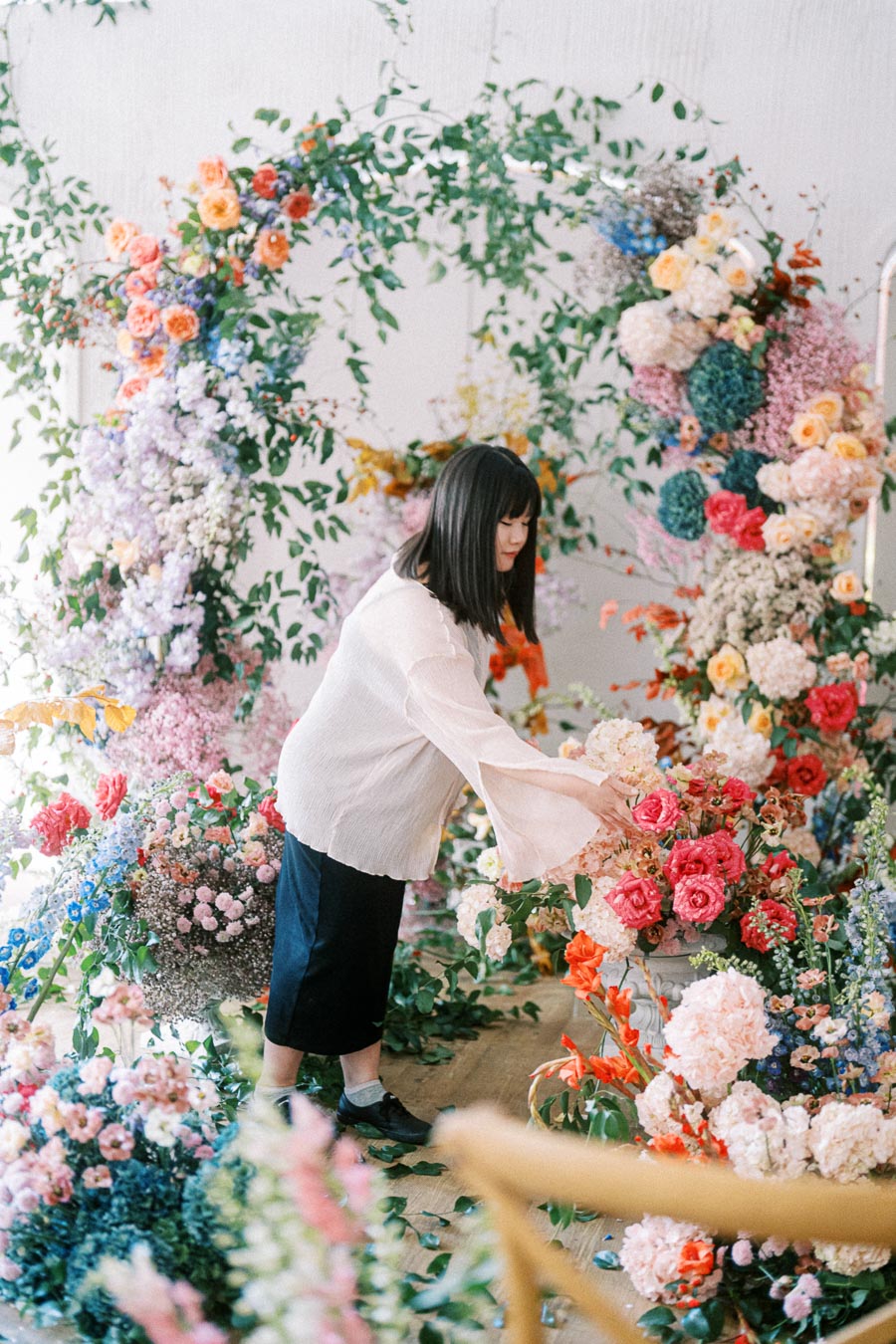 Woman arranging colorful floral display at an indoor garden event with a variety of flowers including roses and hydrangeas, creating a vibrant and artistic atmosphere.