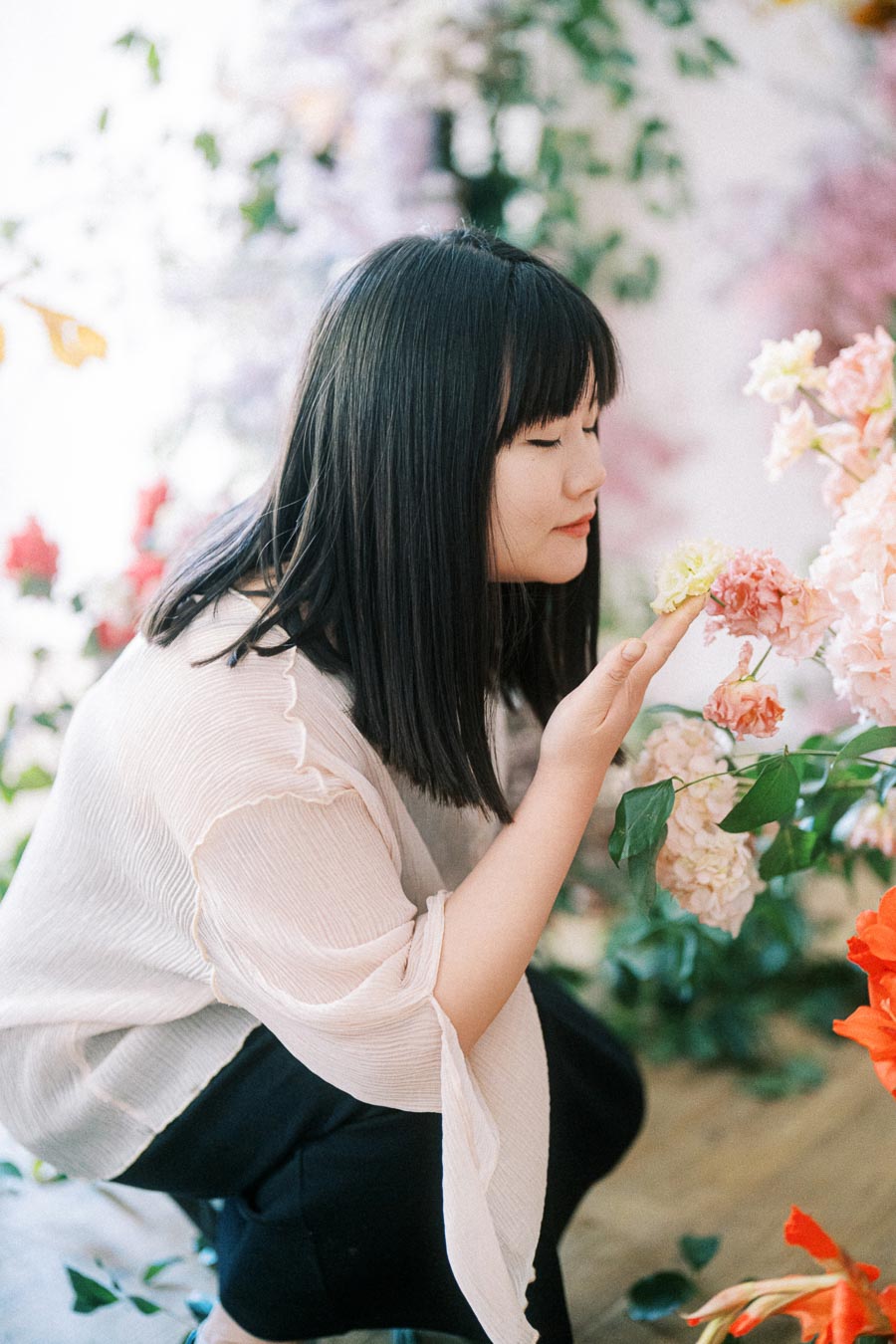 Young woman in a light blouse gently sniffing colorful flowers amidst a lush floral backdrop, conveying a serene and peaceful moment in nature.
