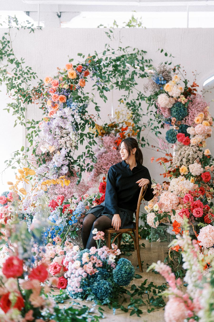 Woman seated surrounded by vibrant floral arrangements featuring roses, hydrangeas, and greenery, creating a lush, colorful backdrop in a bright setting.