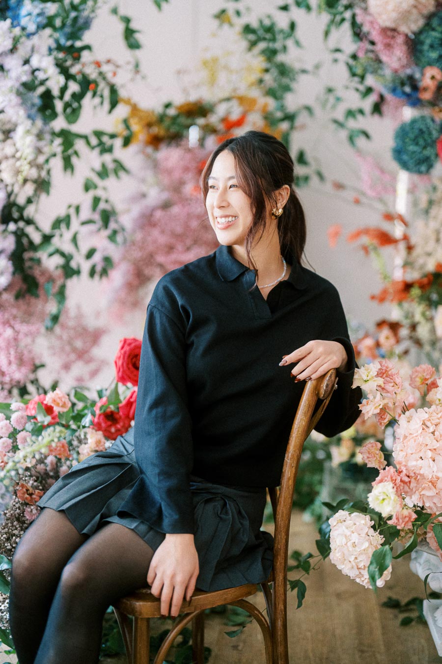 A person sitting on a wooden chair surrounded by vibrant and colorful flowers, wearing a black top and skirt, with a cheerful expression and natural lighting highlighting the floral backdrop.