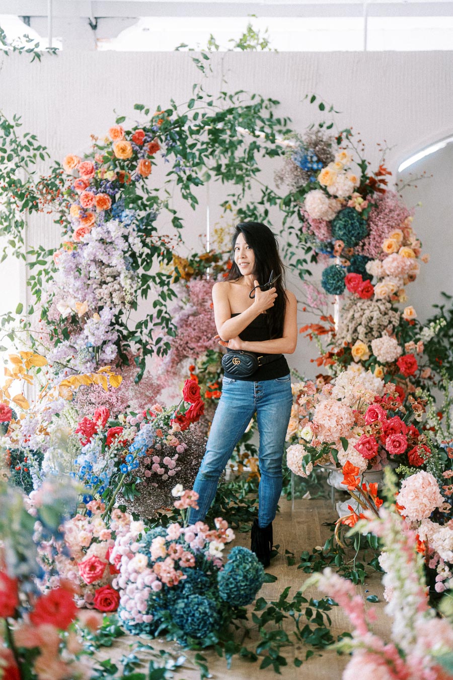 A florist standing amidst a vibrant floral installation with multicolored roses, hydrangeas, and greenery, holding pruning shears and smiling.