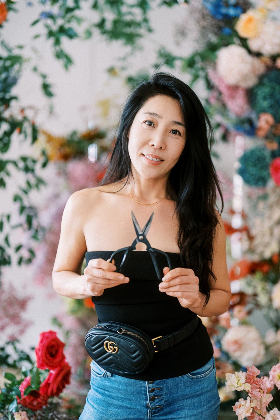 A woman standing in a floral shop, holding pruning scissors, dressed in a black top and jeans, with a stylish black belt bag.