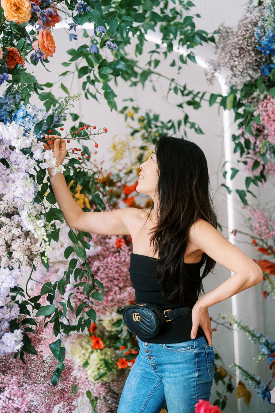 A woman in a black top and jeans enjoys a vibrant floral display, surrounded by colorful flowers and greenery.