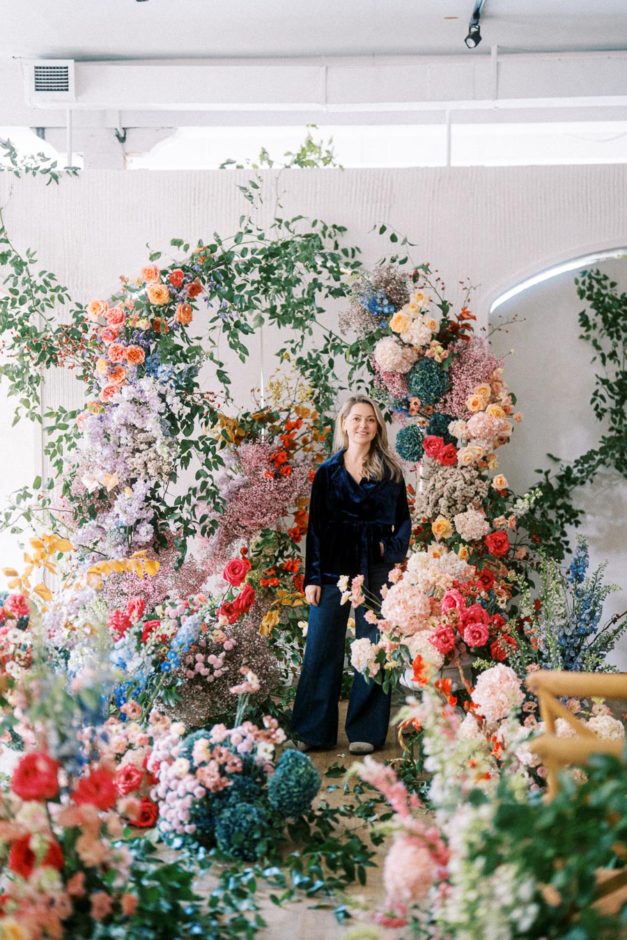 A person stands amidst a vibrant floral display featuring an array of colorful flowers and greenery, arranged artistically against a white backdrop.