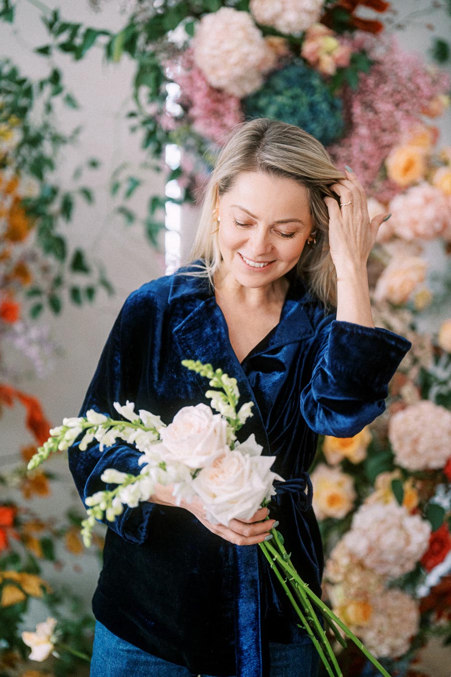 Woman in a blue velvet shirt smiling while holding a bouquet of roses and snapdragons, surrounded by a vibrant floral background.