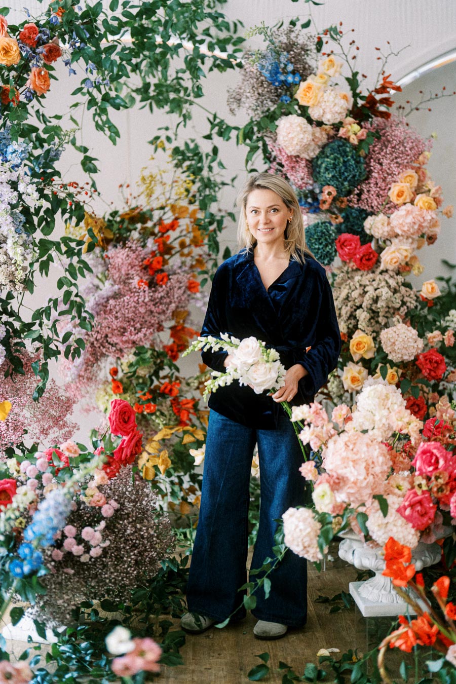 A person standing amidst a vibrant array of colorful flowers and lush greenery, holding a bouquet. The setting conveys a sense of creativity and passion for floral design.