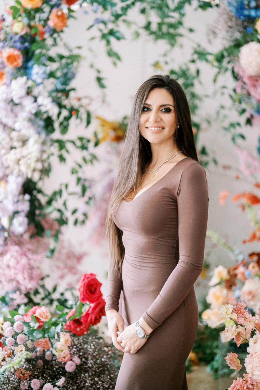 A woman in a brown dress stands smiling in front of a colorful floral backdrop, featuring a variety of vibrant flowers and greenery.