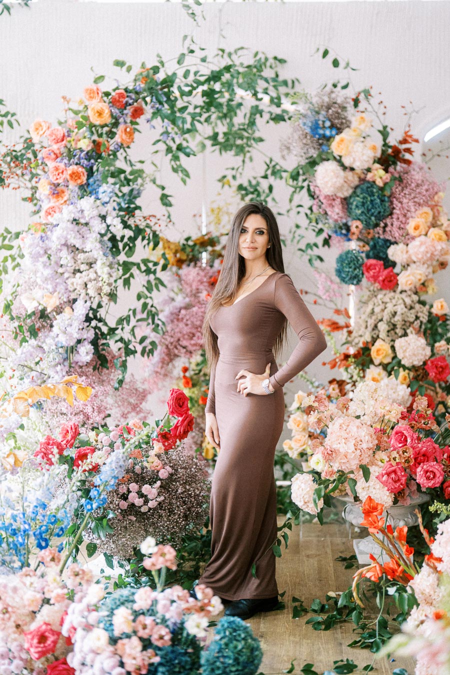 A woman in a long brown dress stands surrounded by a vibrant floral arrangement, featuring red, pink, orange, and blue flowers, creating an elegant and colorful atmosphere.