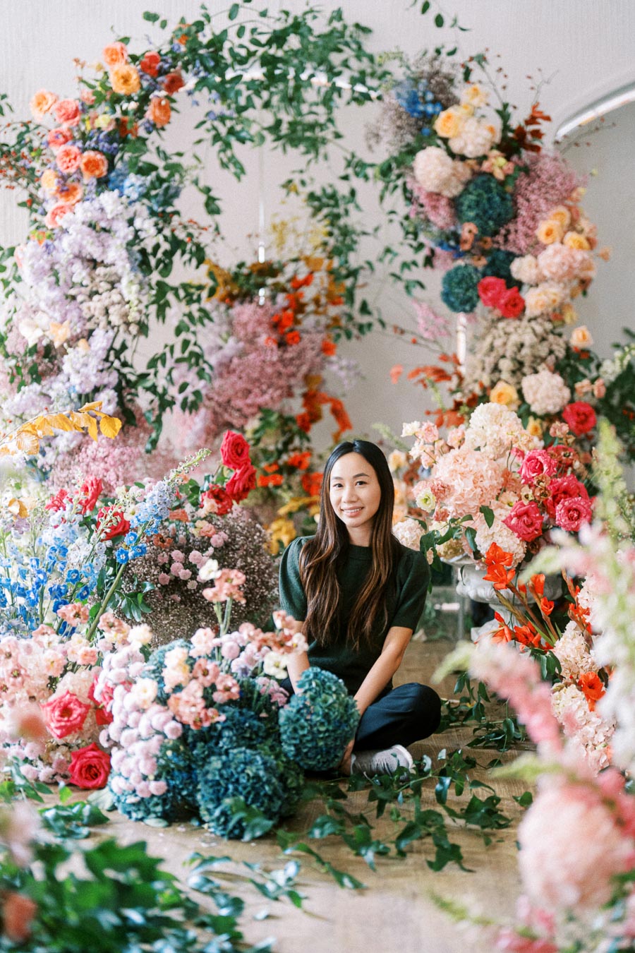A woman surrounded by a vibrant floral arrangement, featuring an array of colorful flowers such as roses, hydrangeas, and greenery, creating a lush and artistic setting.