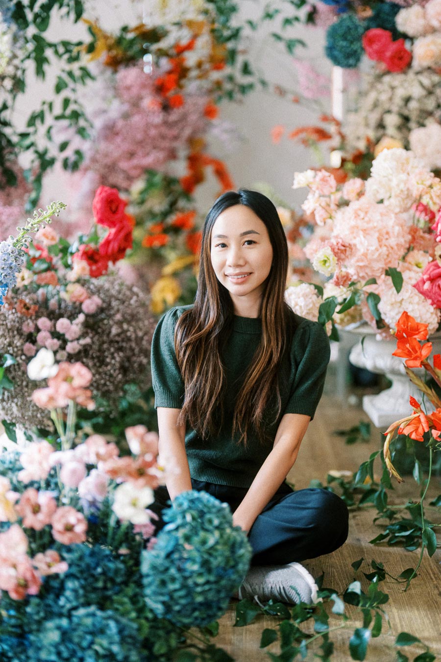 A person sitting on the floor surrounded by a vibrant array of colorful flowers, wearing a dark green sweater and smiling. The floral arrangement includes pink, red, blue, and orange blooms, creating a lush and artistic backdrop.