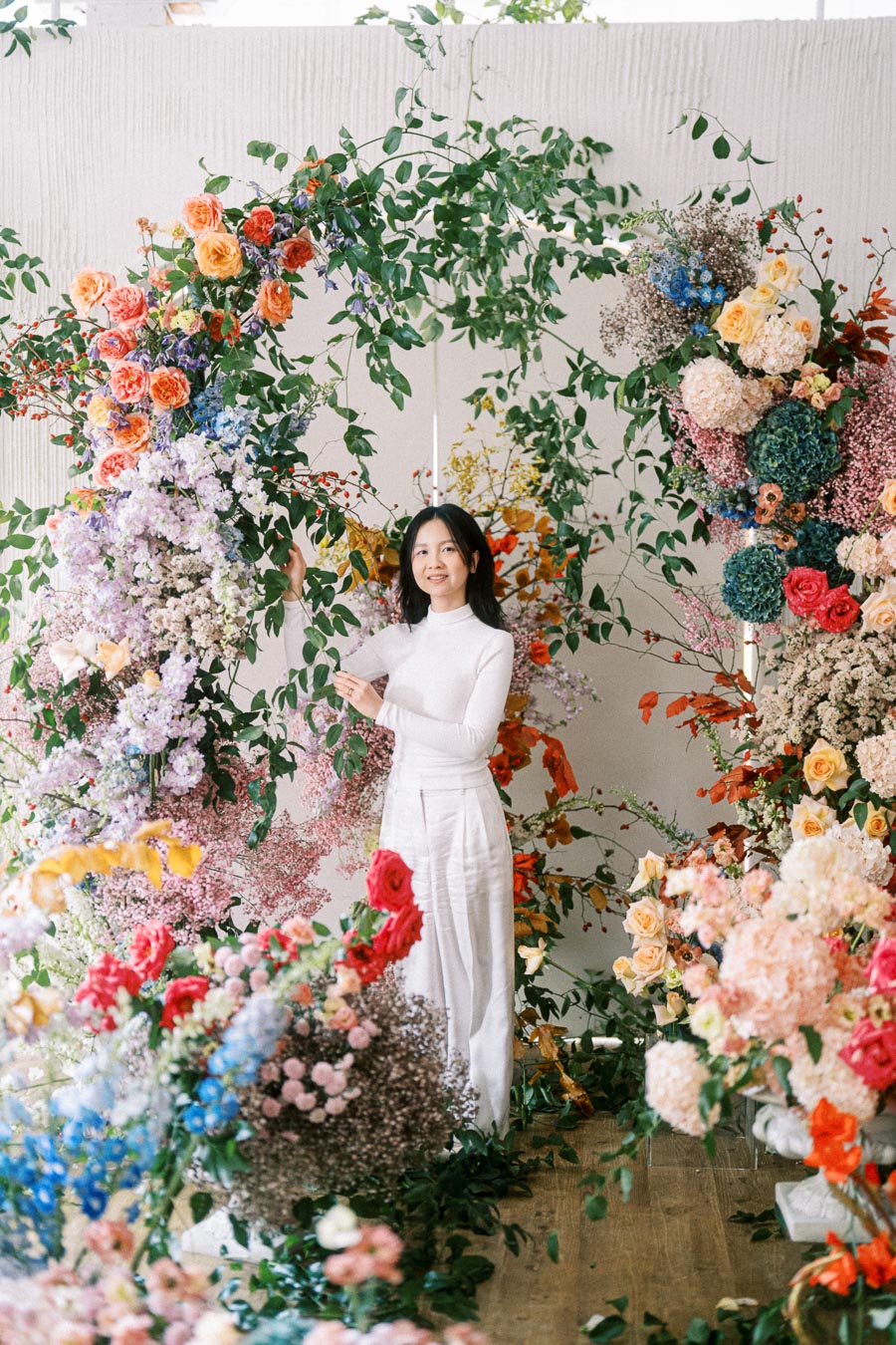 A woman in a white outfit stands amidst a vibrant floral arrangement with various colorful flowers and lush greenery, set against a neutral background.