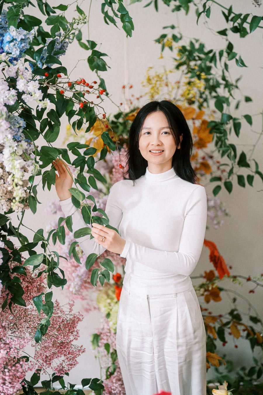A person in a white outfit standing amid lush greenery and colorful flowers, smiling gently. The scene captures a serene and natural ambiance, highlighting the beauty of floral arrangements.