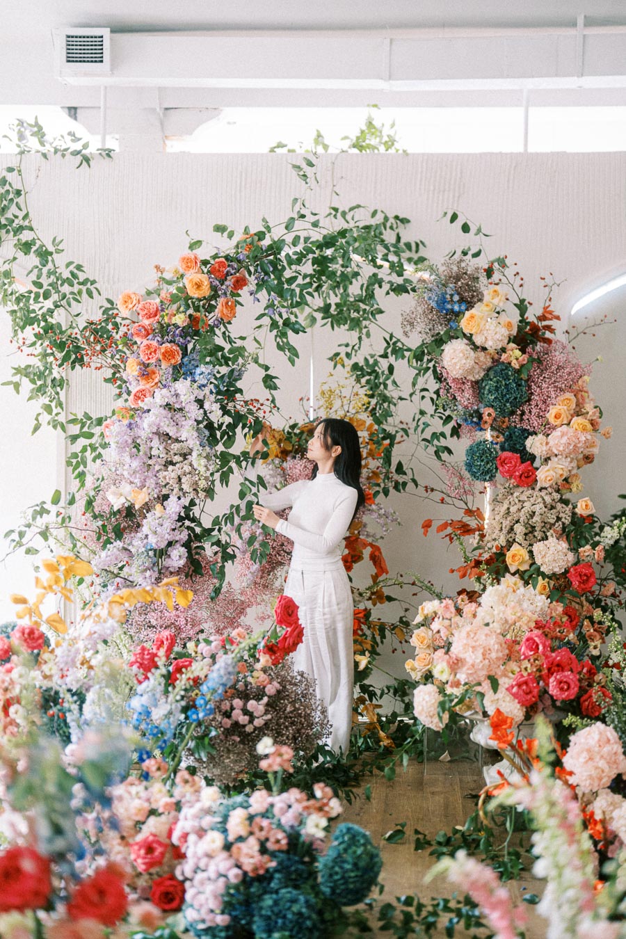 A woman in white arranging a vibrant floral display with various colorful flowers and green leaves inside a bright, airy space.