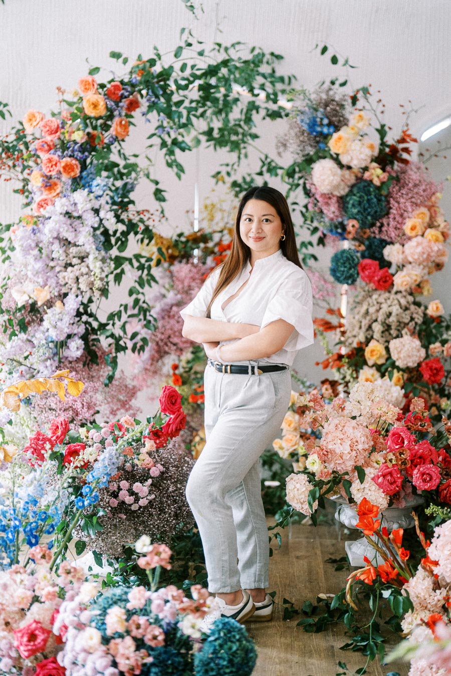 A woman in a white blouse and light gray pants stands confidently surrounded by a vibrant floral arrangement featuring a variety of colorful flowers like roses and hydrangeas, creating a lush and artistic backdrop.