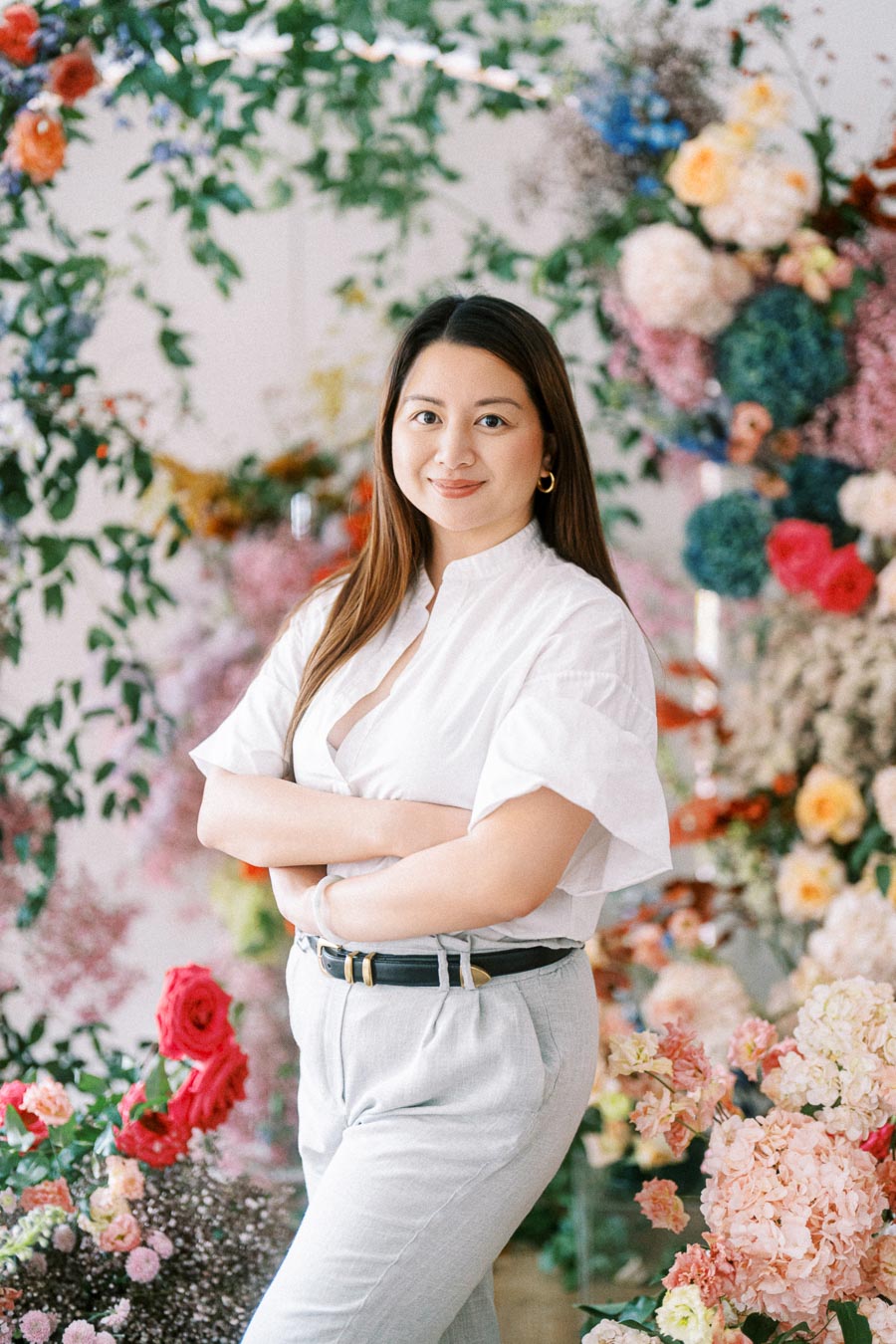 Portrait of a confident woman standing with arms crossed amidst a vibrant floral arrangement, wearing a white shirt and light gray pants. The background features lush greenery and colorful flowers, creating an inviting and creative atmosphere.