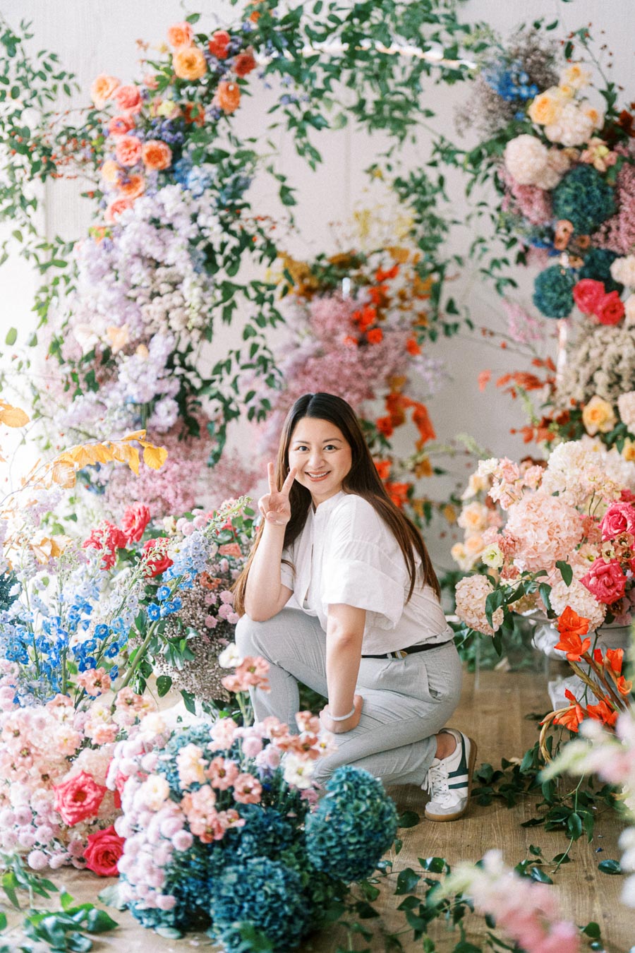 Young woman posing in a colorful flower arrangement display, showcasing vibrant blooms and greenery indoors, with a cheerful expression and peace sign.