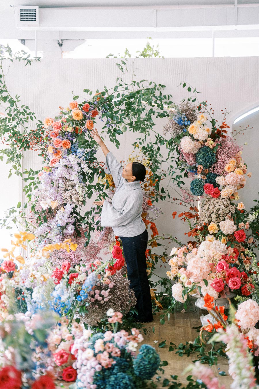 A person arranging a colorful floral display with a variety of blooms including roses, hydrangeas, and greenery, creating an arch in a bright, indoor setting.