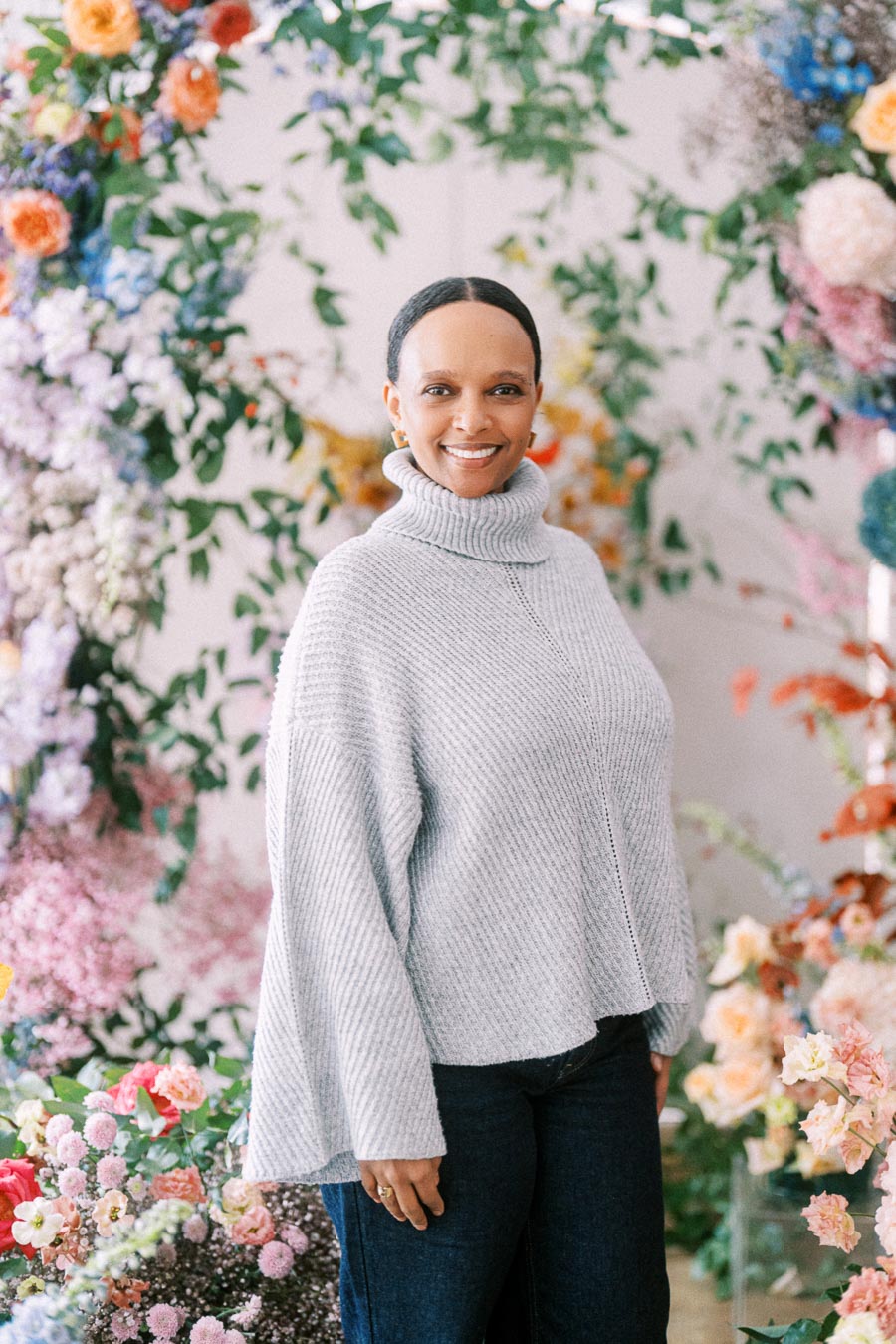 A person wearing a gray sweater smiles amidst a vibrant floral background with various colorful flowers and greenery.