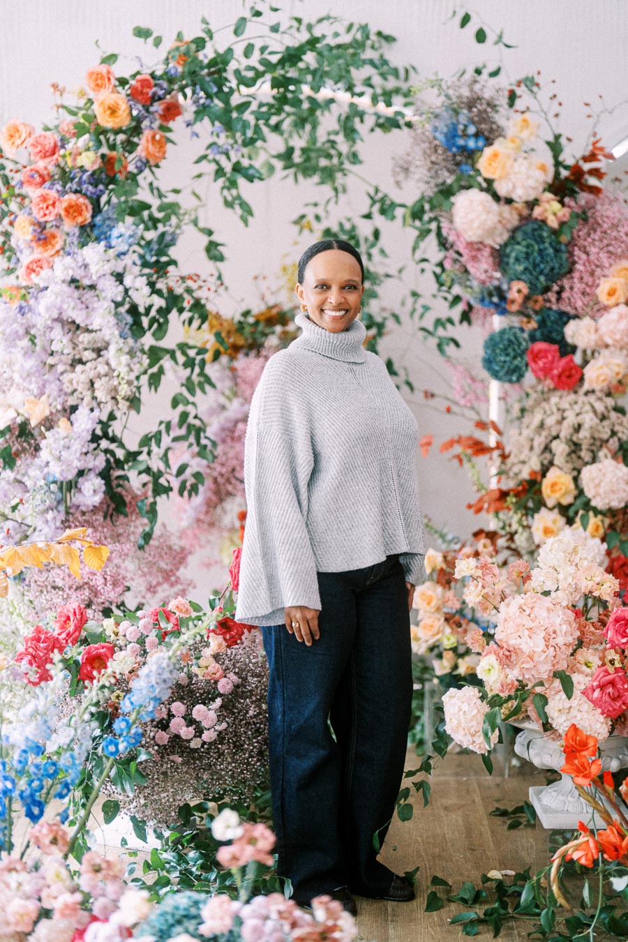 A woman wearing a gray sweater stands in front of a vibrant floral display with a variety of colorful flowers and greenery.
