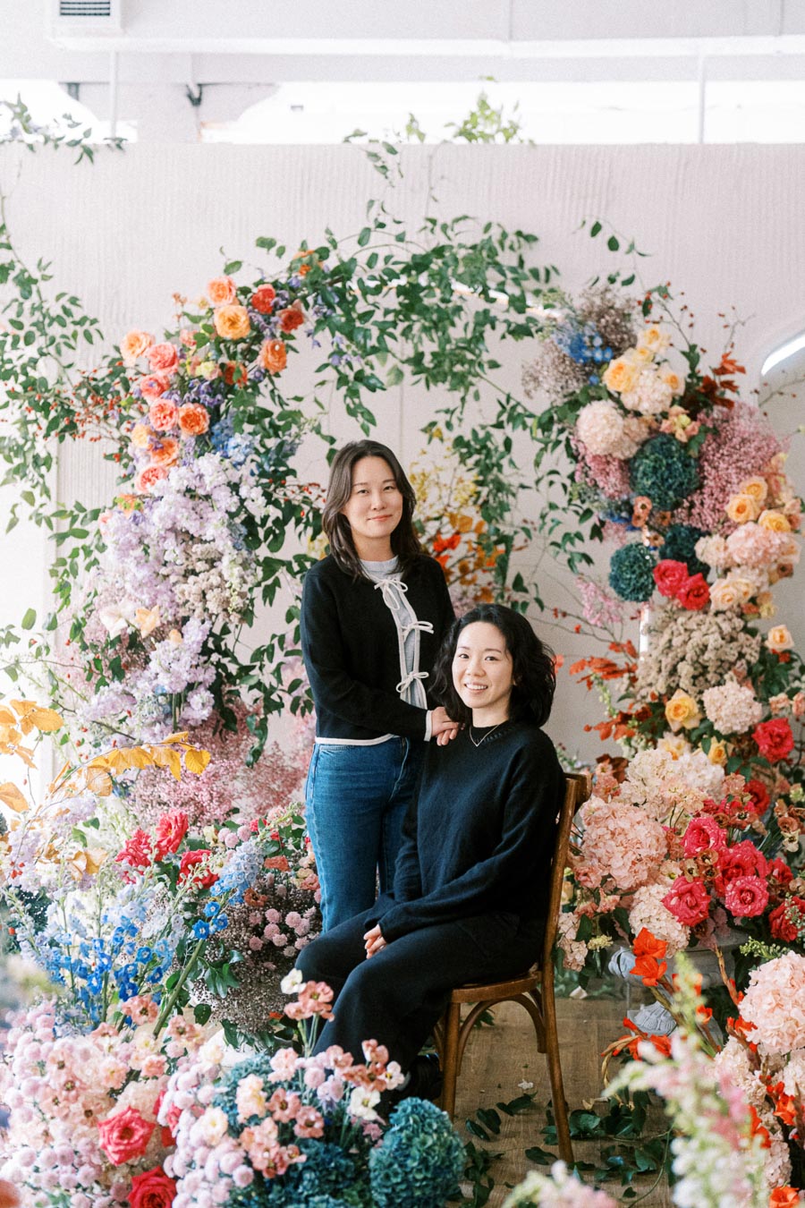 Two women surrounded by vibrant, colorful floral arrangements, standing and sitting against a backdrop of lush greenery and assorted flowers, creating a visually stunning garden-like scene indoors.